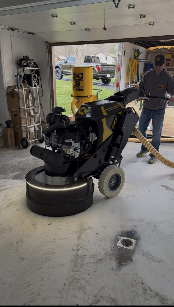 Epoxy King operator using a SASE concrete floor grinder connected to dust-extraction equipment in a garage